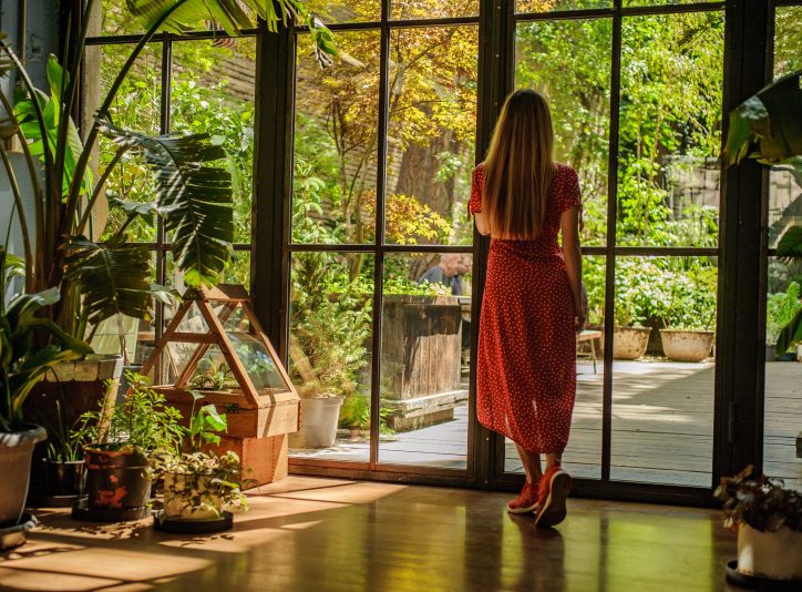 Cute girl in beautiful red dress looking at the large window in doors